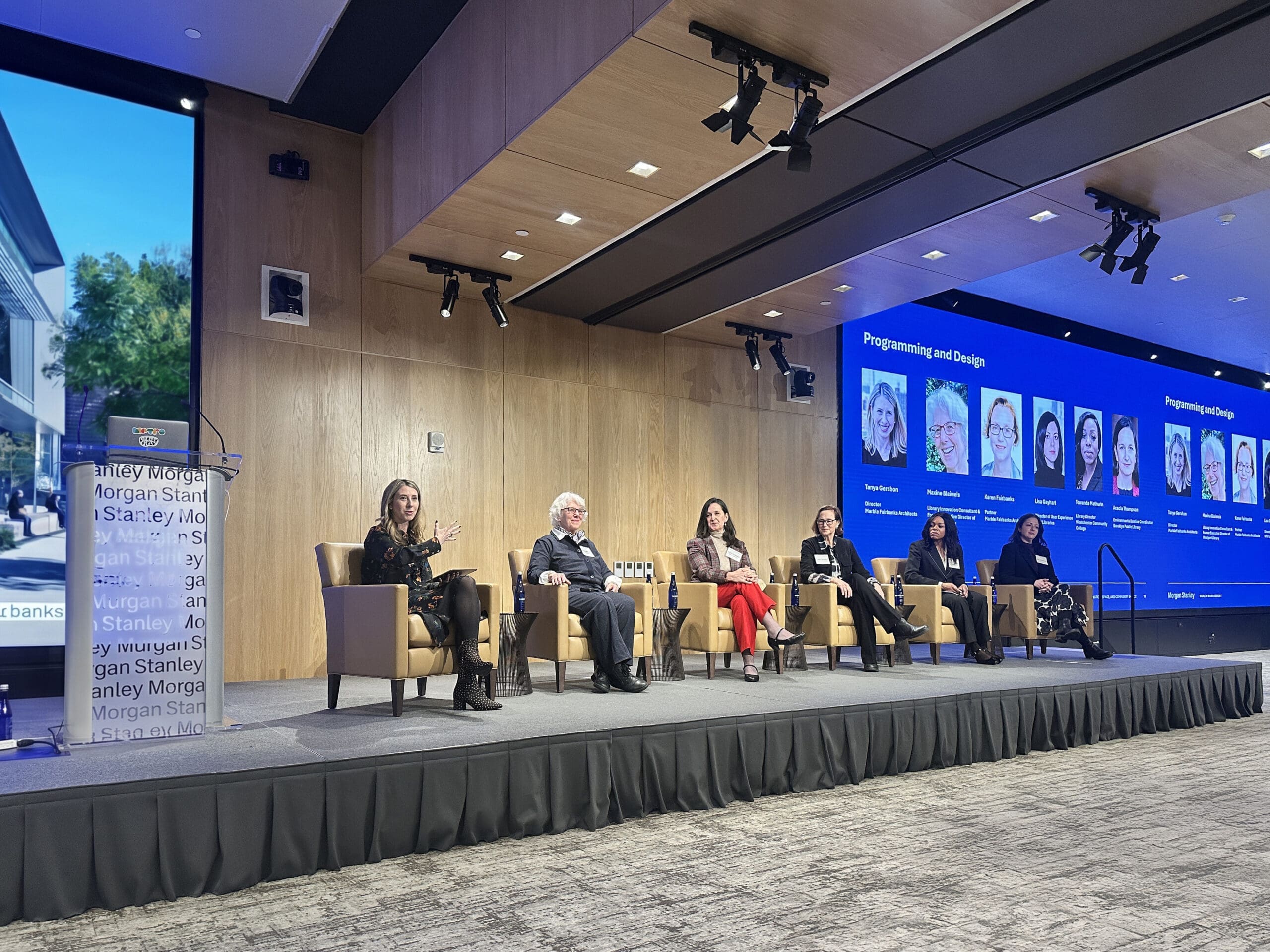 Six panelists seated in armchairs on stage at Reimagining Libraries Programming and Design panel, with one speaking.